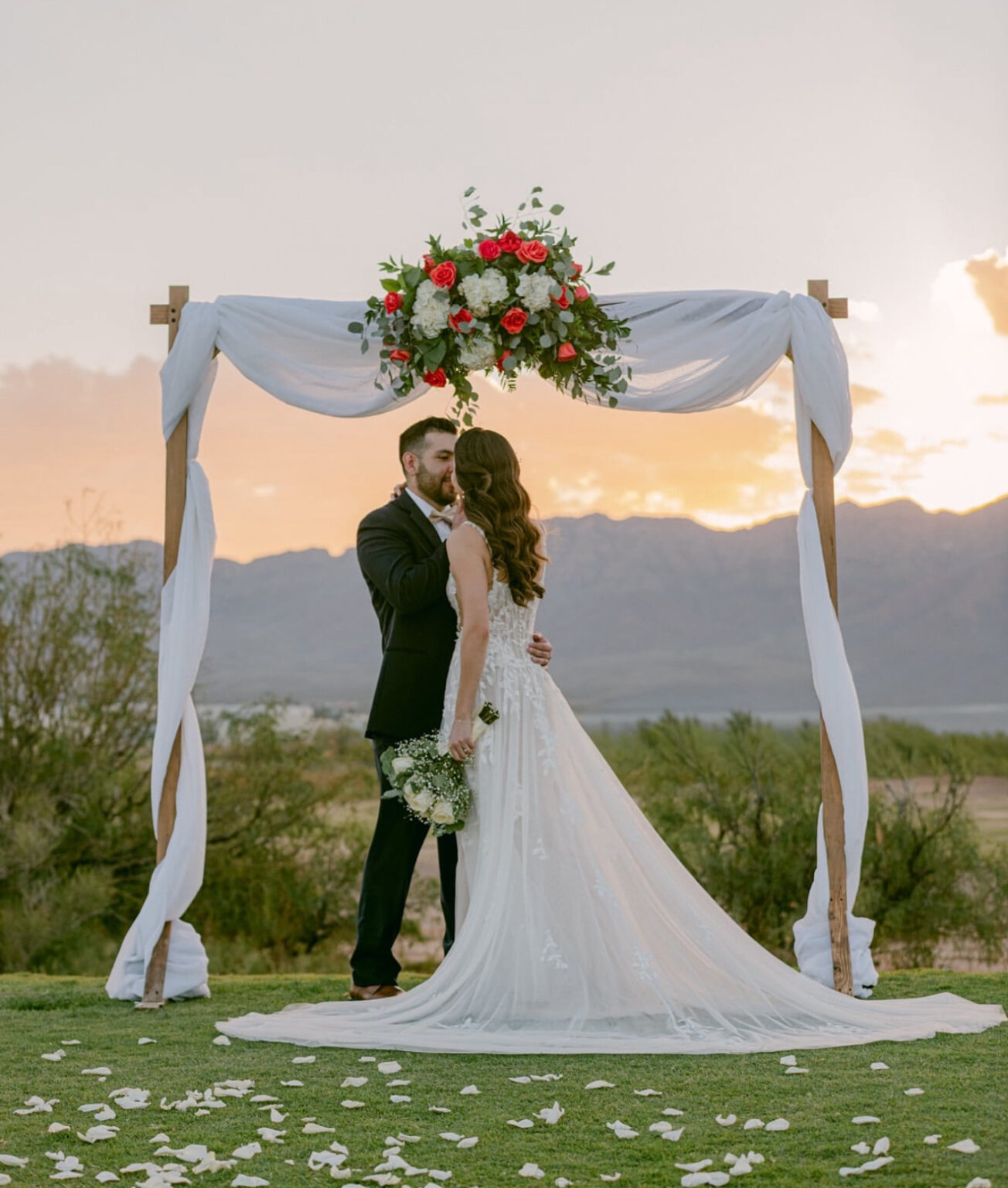 Bride and groom share an intimate moment beneath a floral wedding arch during a sunset ceremony at Painted Dunes Desert Golf Course in El Paso, with mountain views and rose petals creating a romantic outdoor wedding setting.