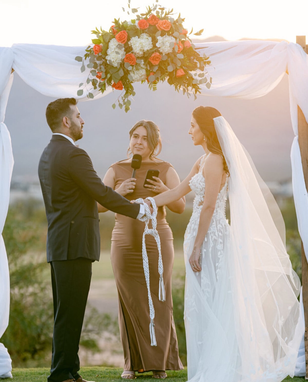 Bride and groom participate in a unity ritual during their outdoor wedding ceremony at Painted Dunes Desert Golf Course in El Paso, framed by a floral arch, desert scenery, and soft sunset lighting over the Franklin Mountains.