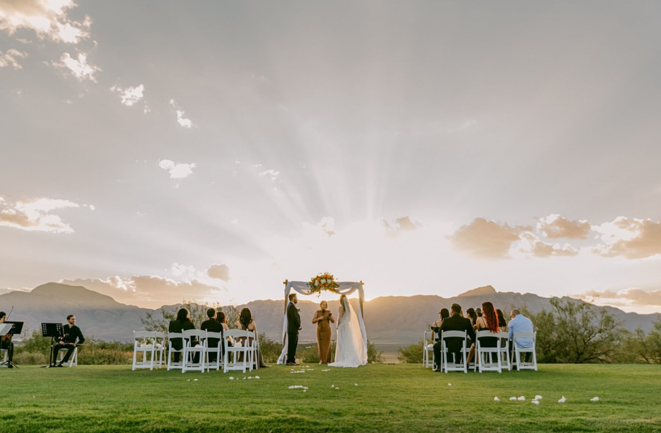 Wide-angle view of an outdoor wedding ceremony at Painted Dunes Desert Golf Course in El Paso, with the bride and groom standing beneath a floral arch as dramatic sunset rays shine over the Franklin Mountains.