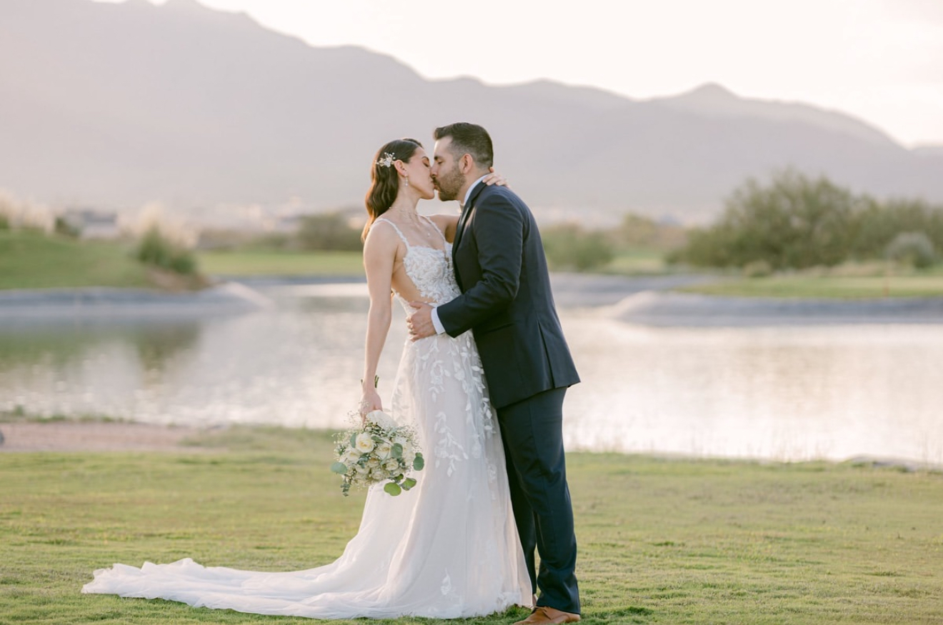 Bride and groom share a romantic kiss beside the lake at Painted Dunes Desert Golf Course in El Paso, with mountain views and soft sunset lighting creating a stunning outdoor wedding moment.